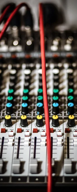 A detailed view of an audio mixing desk with colorful knobs, sliders, and red cables connected. A detailed view of an audio mixing desk with colorful knobs, sliders, and red cables connected.